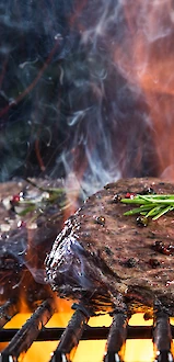 Grilled steak on a barbecue with flames, seared crust, and a sprig of rosemary on top.