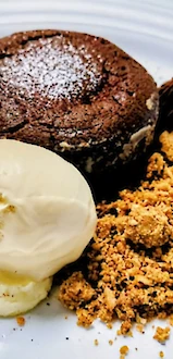 A chocolate fondant cake with vanilla ice cream, a dusting of powdered sugar, chocolate decorations, and a crumbly crumble on the side.