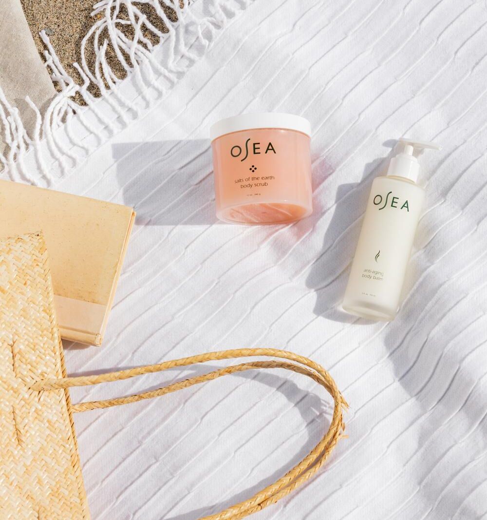 A beach scene with a white striped towel, a peach OSEA jar, a white OSEA bottle, a sponge, a beige bag, and a straw tote on sand, sunlit and calm.