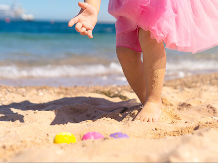 A child's feet in the sand at the beach, pink tutu visible, colorful plastic toys nearby, ocean waves and a boat in the distance. End.