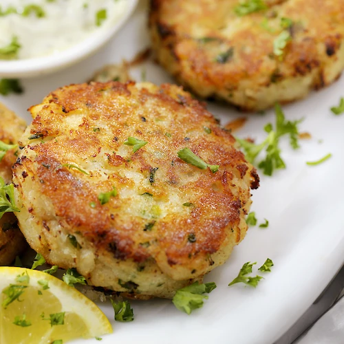 Delicious-looking golden brown potato or herb patties with herbs, lemon wedges, and a dipping sauce on the side, perfect for a fresh bite.