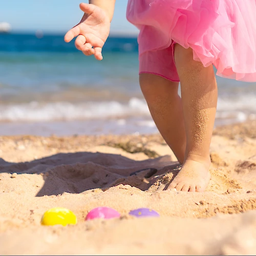 A child in a pink dress is playing in the sand on the beach, with colorful shells or toys near the feet and the sea in the background.