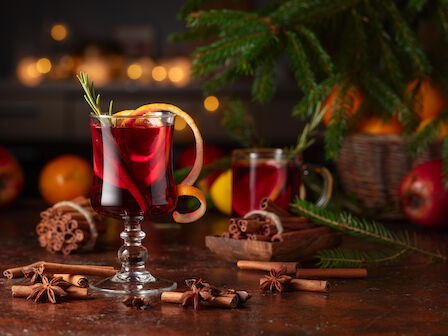 Two glasses of red mulled wine with citrus peels, surrounded by cinnamon sticks, star anise, pine branches, and seasonal fruits on a festive table.