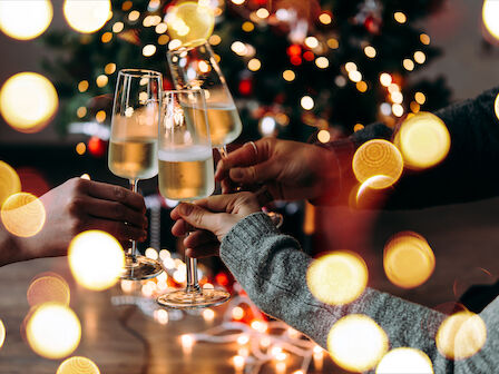 People cheers with champagne glasses in a festive, bokeh-lit celebration near a Christmas tree.