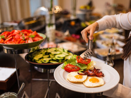 A person serving a plate from a buffet with sunny-side eggs, tomatoes, and grilled vegetables, surrounded by more dishes.