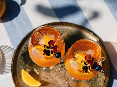 Two orange cocktails with flowers and blueberries on a tray, sliced oranges nearby, outdoors on a striped tablecloth.