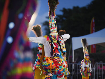 Colorful performers in tall, decorated costumes and headdresses, dancing at an outdoor festival.