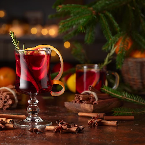 Two glass mugs with red drinks on a festive table, surrounded by cinnamon sticks, star anise, oranges, pine branches, and cookies, cozy holiday setup.