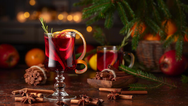 Two glass mugs with red drinks on a festive table, surrounded by cinnamon sticks, star anise, oranges, pine branches, and cookies, cozy holiday setup.