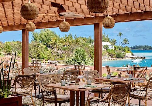 A seaside dining area with wicker chairs, wooden tables, and hanging lanterns under a tiled roof, overlooking turquoise water and green shrubbery.
