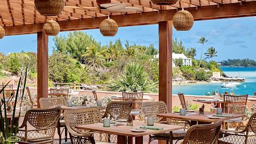 A seaside dining area with wicker chairs, wooden tables, and hanging lanterns under a tiled roof, overlooking turquoise water and green shrubbery.