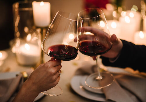Two people clink wine glasses at a candlelit table, toasting a romantic dinner with dim, warm lighting and blurred background.