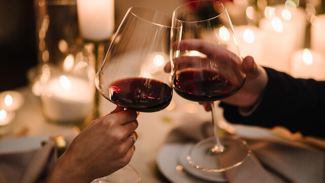 Two people clink wine glasses at a candlelit table, toasting a romantic dinner with dim, warm lighting and blurred background.
