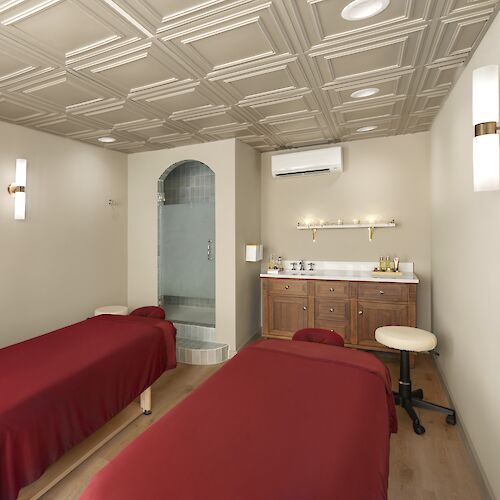 Two spa beds with burgundy coverings in a small treatment room, a wooden cabinet with a mirror, and soft lighting.