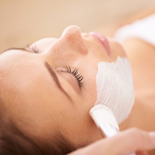 A woman lying down with a white facial mask being applied, eyes closed, relaxing spa treatment.