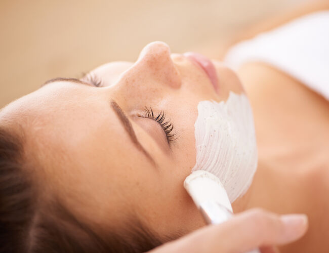 A woman lying down with a white facial mask being applied, eyes closed, relaxing spa treatment.