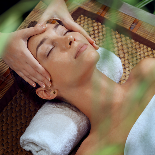 A woman enjoys a peaceful facial massage outdoors, eyes closed, head resting on a towel, while hands work gently on her temples.