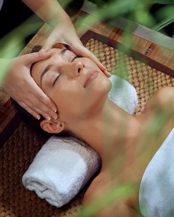 A woman enjoys a peaceful facial massage outdoors, eyes closed, head resting on a towel, while hands work gently on her temples.