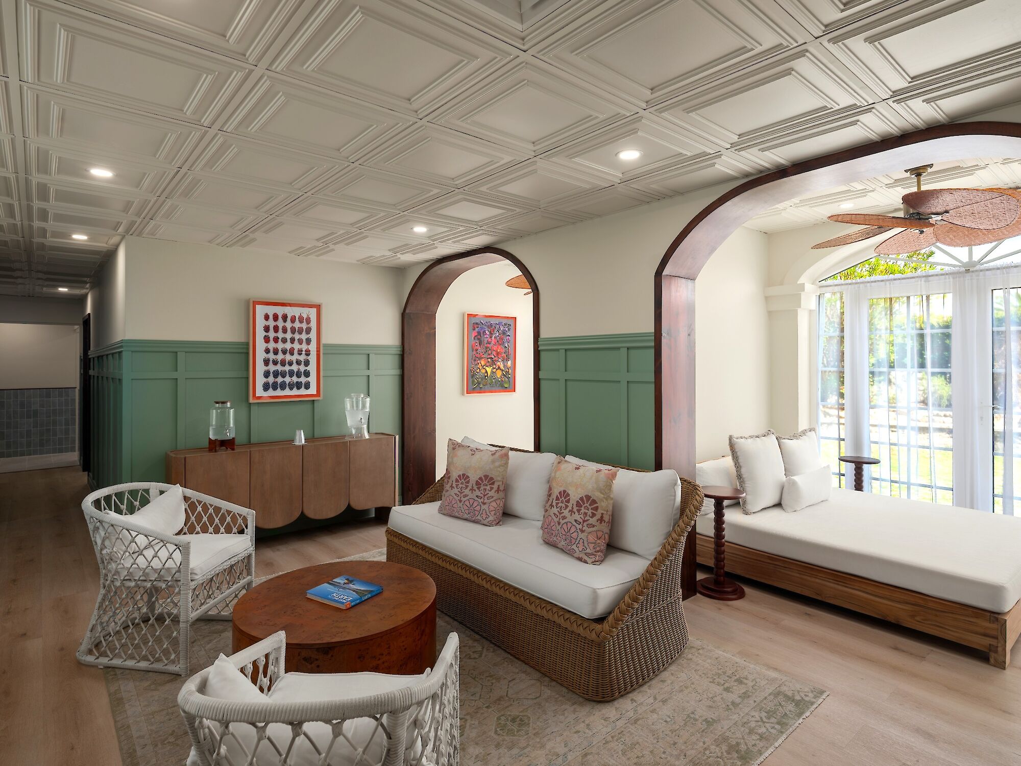 A bright, airy lounge with a wicker sofa and chairs, a round wooden coffee table, built-in daybed under arches, green wainscoting, and ceiling fans.