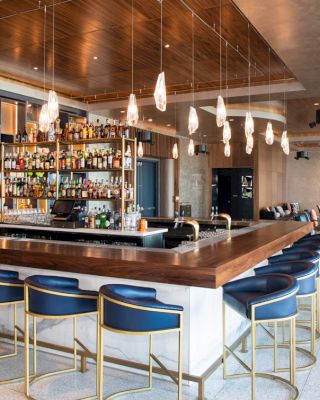 A modern bar with a wooden counter, blue chairs, pendant lights, and a stocked back bar. There's a seating area in the background and a sleek design.