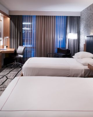 The image shows a modern hotel room with two neatly made beds, a TV on the wall, a desk with a chair, and ambient lighting with a stylish decor.