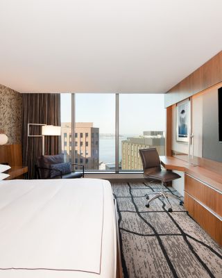 The image shows a modern hotel room with a large bed, TV, desk, chair, and a view of buildings and water through a floor-to-ceiling window.