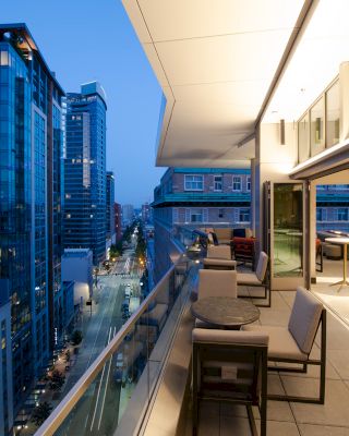 The image shows a modern cityscape with tall buildings, viewed from a balcony of a sleek apartment or office space with outdoor seating.
