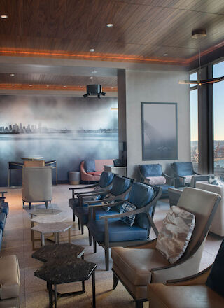 A modern lounge with comfortable seating, large windows showcasing a waterfront view, and contemporary decor including a cityscape mural on the wall.
