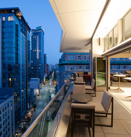 A modern balcony with seating and tables overlooks an urban cityscape with high-rise buildings and a street below, at dusk or early evening.