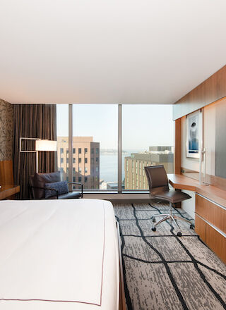 A modern hotel room with a large bed, desk, chair, flat-screen TV, and a window with a city view.