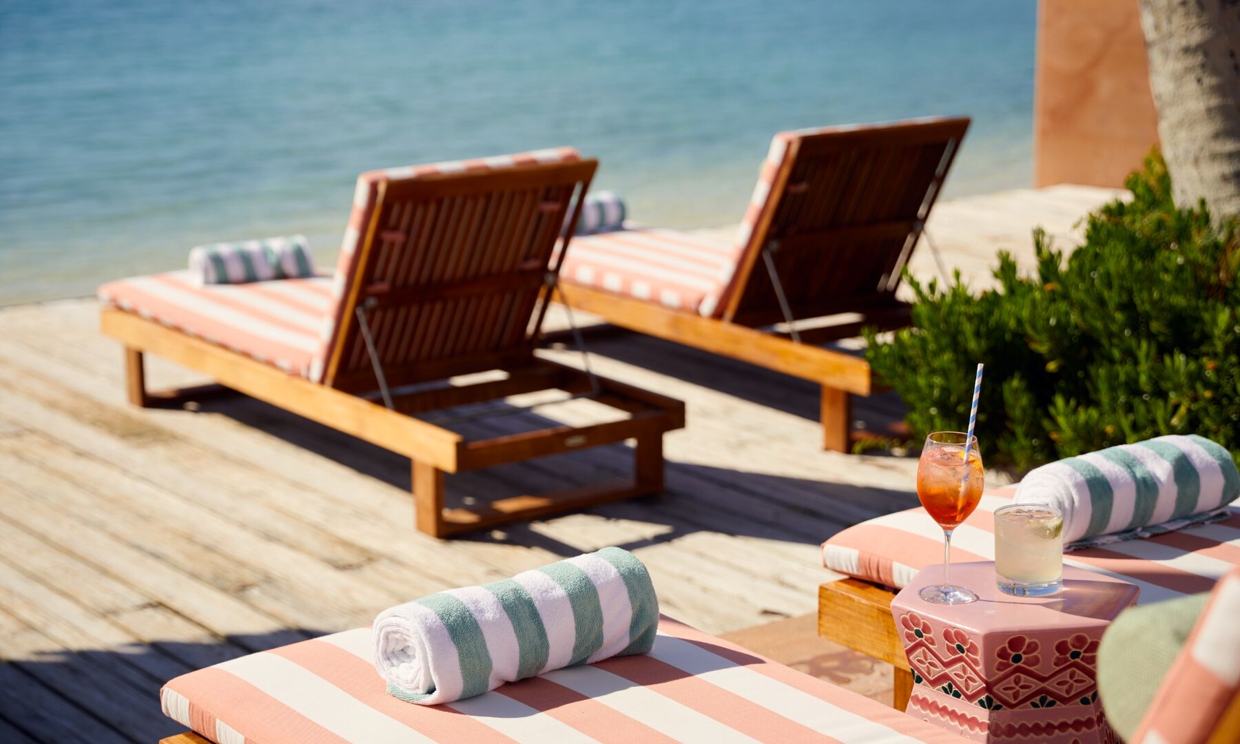 Beachside lounge setup with striped sunbeds, rolled towels, and tropical drinks on a wooden deck by clear blue water.