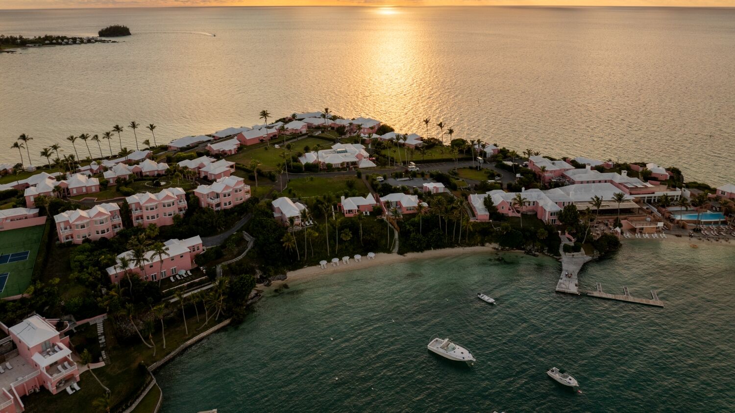 Haloed coastal resort at sunset with villas along a curved shoreline, calm turquoise water, and small boats anchored near the shore.