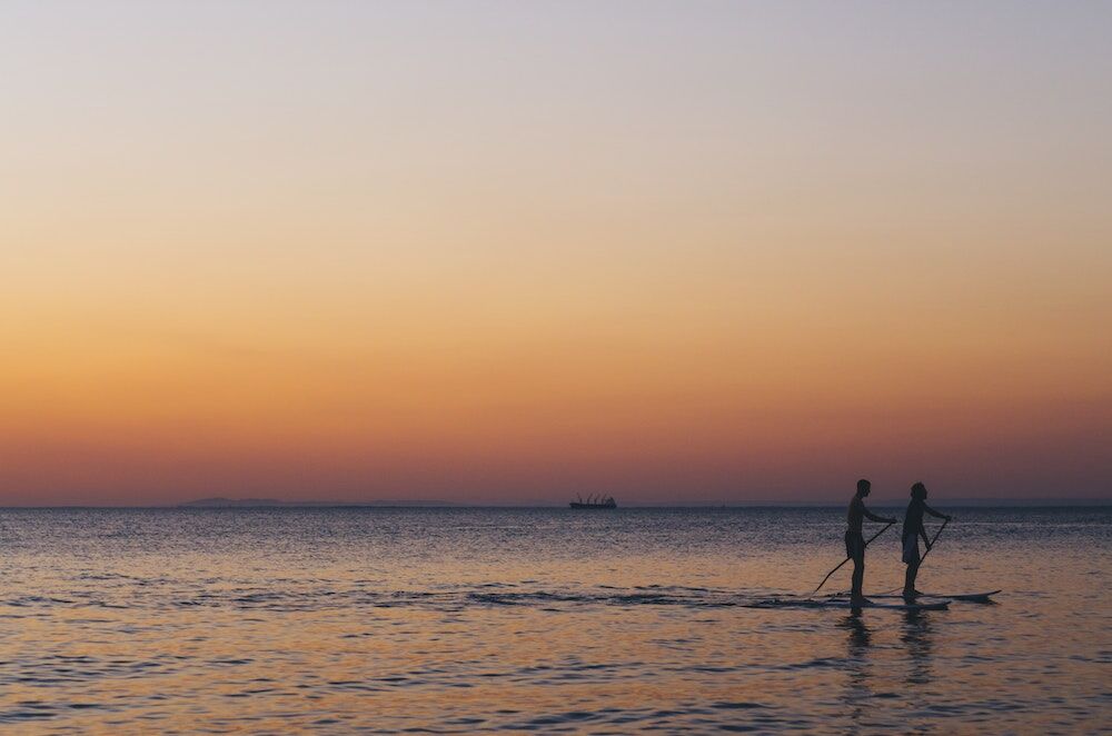 Two people are paddleboarding on calm water during a serene sunset, with a distant ship silhouette on the horizon.