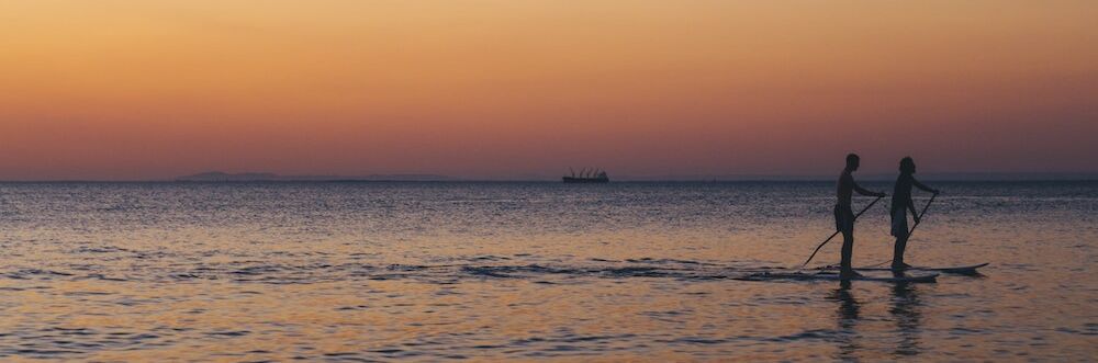 Two people are paddleboarding on calm water during a serene sunset, with a distant ship silhouette on the horizon.