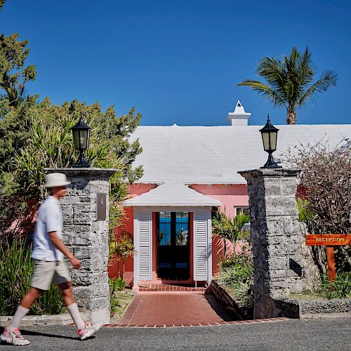 A person walks past an entrance with stone pillars and lamps, leading to a pink building with a white roof, surrounded by greenery and a palm tree.