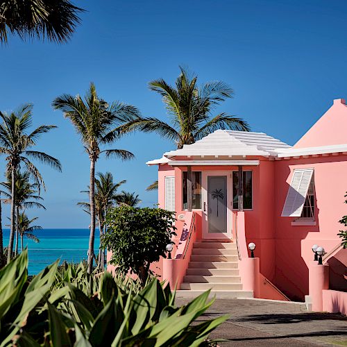 A pink house with white trim is situated near a beach, surrounded by palm trees and lush greenery, with a bright blue sky overhead.