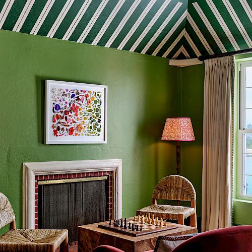 A room with green walls and a patterned ceiling, containing a fireplace, lamp, chairs, a window with curtains, and a chessboard on a table.