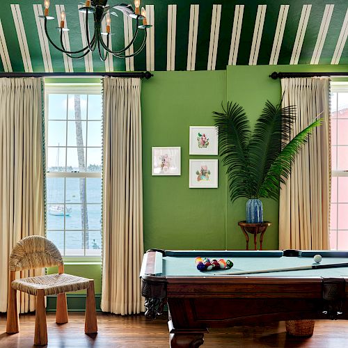 A room with green walls, a pool table, wooden chair, plant, framed pictures, hanging light fixture, and windows with curtains is shown.