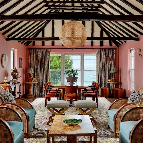 A cozy living room with pink walls, wooden beams, wicker furniture, a central lantern, and large windows allowing natural light to fill the space.