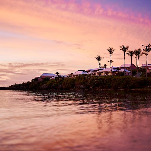 A serene beachside view at sunset, with colorful skies, palm trees, and waterfront houses reflecting on calm waters, creating a tranquil scene.