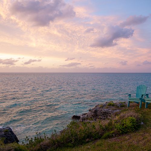 Two turquoise chairs sit on a grassy bluff overlooking a calm ocean under a pastel sunset sky with scattered clouds.
