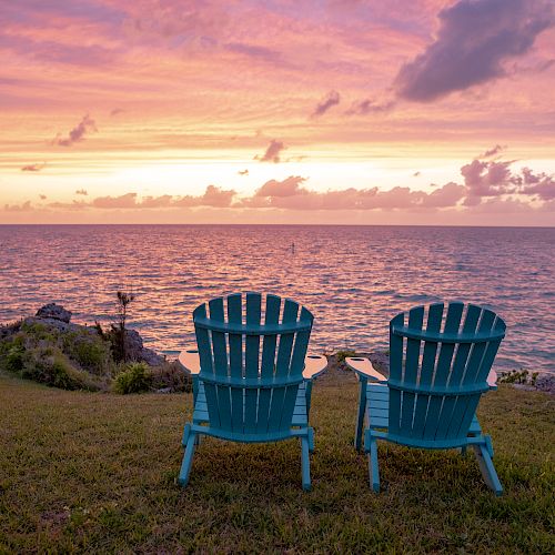 Two blue chairs overlook a serene ocean at sunset, with a vibrant sky filled with orange and pink hues, creating a peaceful and picturesque scene.