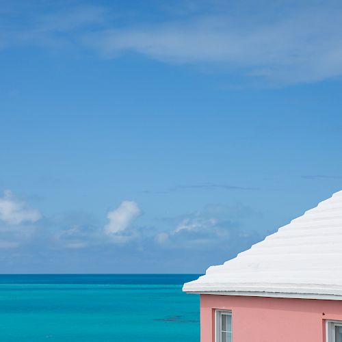 A white-roofed pink building is seen by a vibrant blue ocean under a clear sky, with minimal clouds visible in the background.