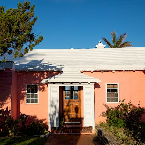 A charming pink house with a white roof and a wooden door, surrounded by greenery and shrubbery, bathed in sunlight.
