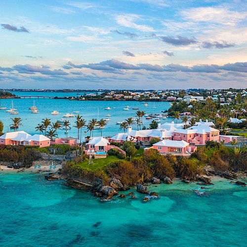 The image shows a coastal scene with vibrant turquoise water, pink houses on a cliff, palm trees, and boats on the water under a partly cloudy sky.